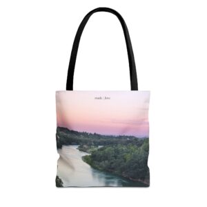 Eco-friendly canvas tote bag with a scenic Sacramento River sunset print, designed by Brenda Nakandi. Features soft golden and pink hues, black straps, and a spacious interior—perfect for nature lovers and everyday use.